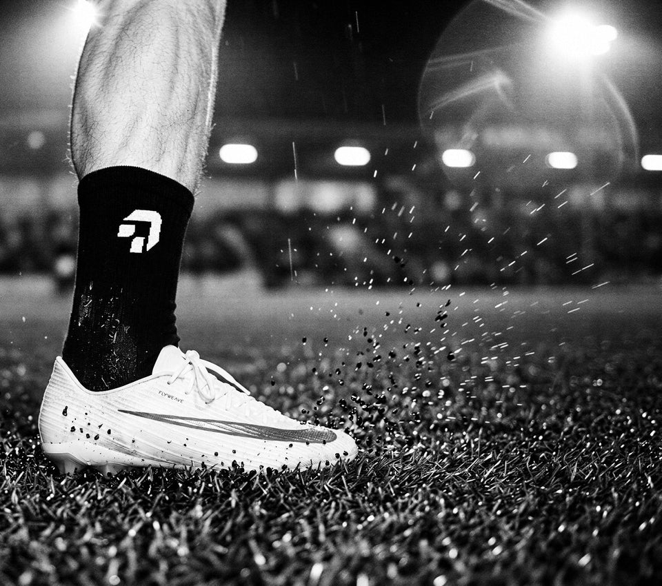 Zwart-wit: close-up van een zwarte sok van Create Performance boven een voetbalschoen op nat kunstgras