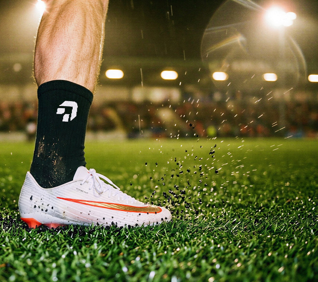 Close-up van een zwarte sok van Create Performance boven een voetbalschoen op nat kunstgras