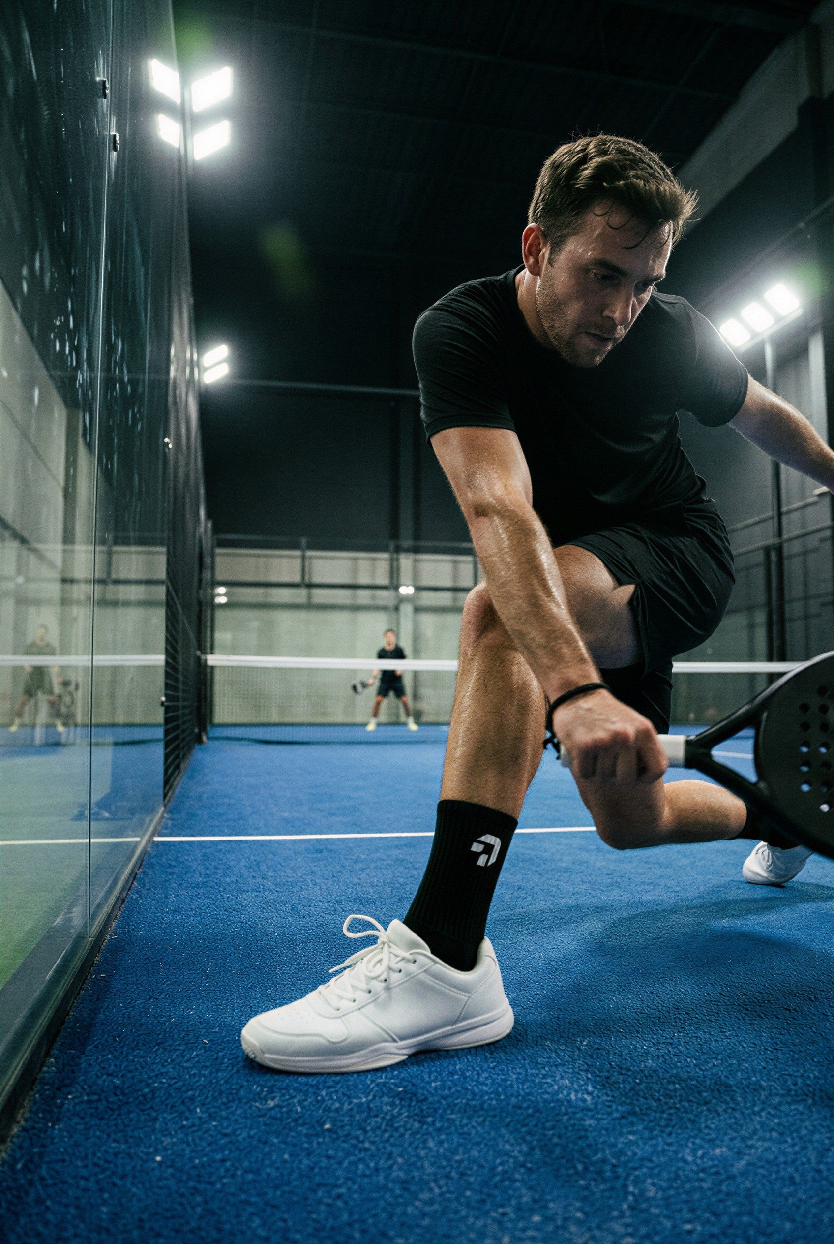 Padelspeler op zwarte gripsokken van Create Performance maakt een diepe uitval naar de bal op een blauwe baan