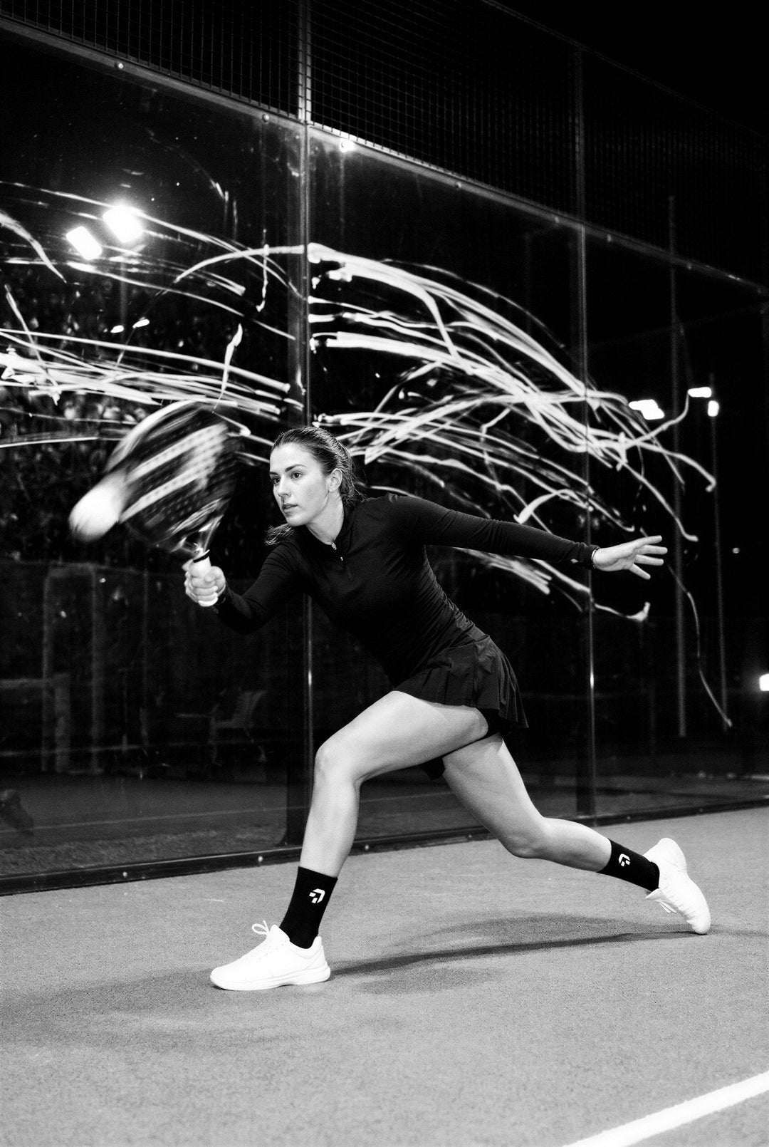 Zwart-wit: padelspeelster op Create Performance sokken duikt naar de bal, lichtsporen van het racket achter haar